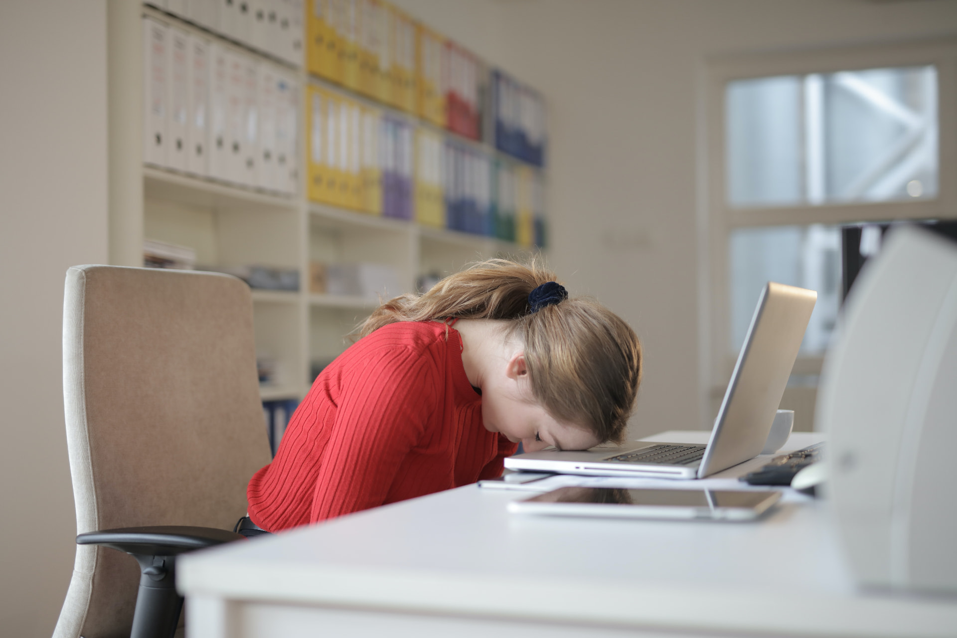 Business owner in a red jumper resting her head on a laptop at a desk, looking overwhelmed